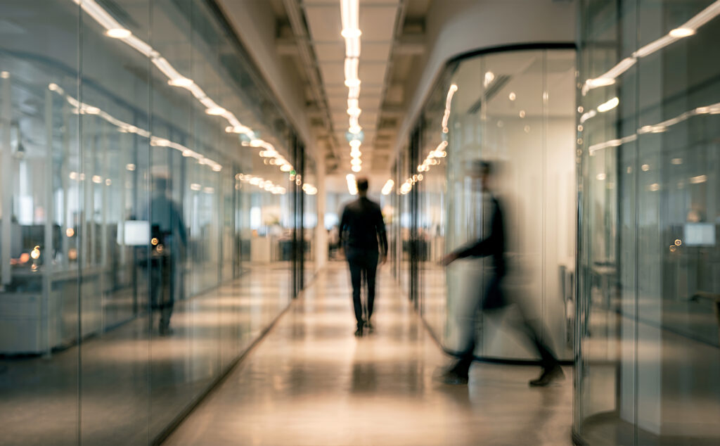 Home a blurred image of professionals walking in a glass walled office corridor, showcasing modern corporate architecture and a busy work environment.